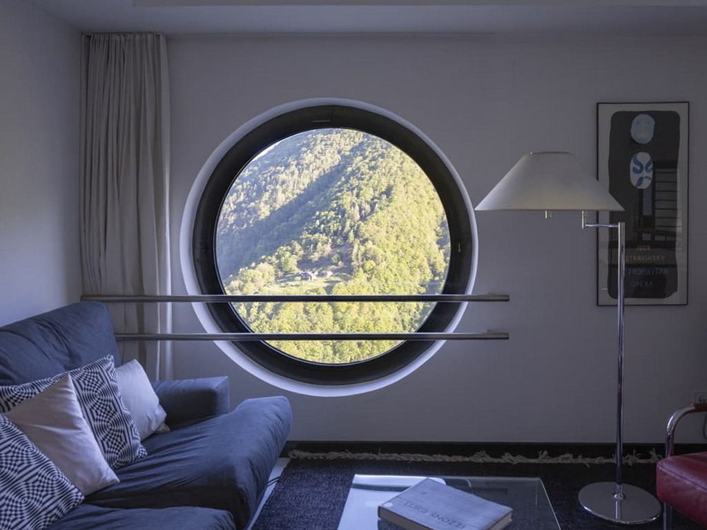Wohnbereich der Casa Walser mit rundem Fenster, Sofa und gerahmtem Blick in die Tessiner Landschaft